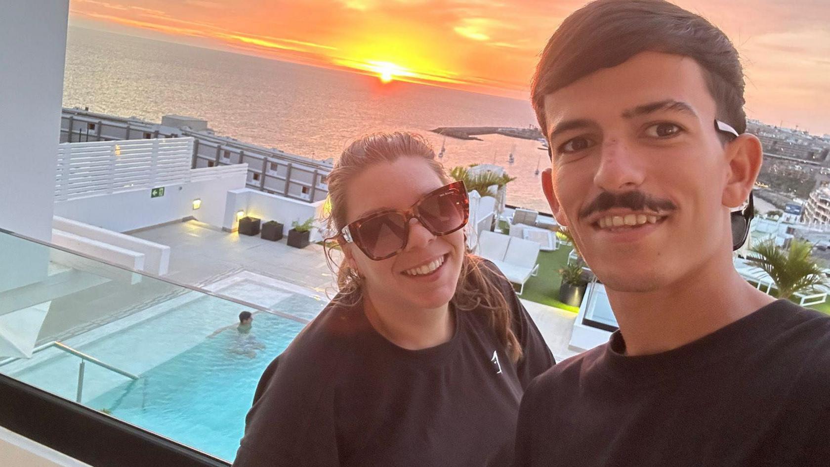 Caprice Betts and her partner Arron Sanchez Garcia standing on a balcony with a swimming pool beneath them and a view out to sea and a setting sun. She is on the left wearing a black T-shirt, has dark brown hair pulled back in a pony tail and is wearing dark glasses. He is on the right with black short hair, a black moustache and is wearing a black T-shirt. They are both smiling. 