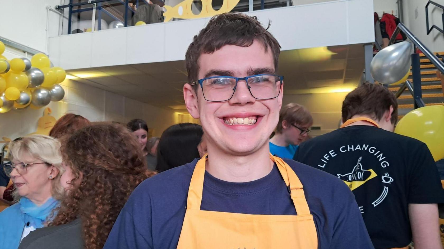 A picture of a young man with brown hair and glasses smiling at the camera. He is wearing a dark blue top and yellow apron, and the busy bakery can be seen behind him.
