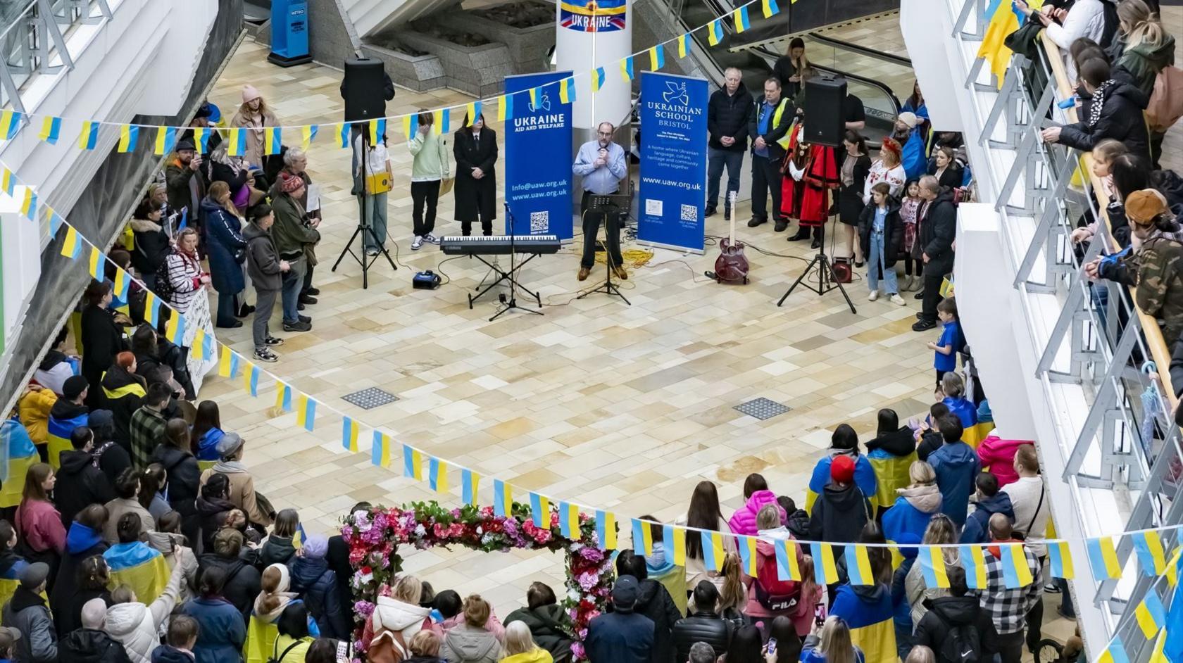 A large group of people gathered around in Broadmead shopping centre. In the middle on the ground floor, there is a man wearing a blue shirt speaking into a microphone. Beside him there is a keyboard on a stand, and behind him there are two large banners which say Ukraine Aid and Welfare. Hanging above them are Ukrainian flags made into bunting. 