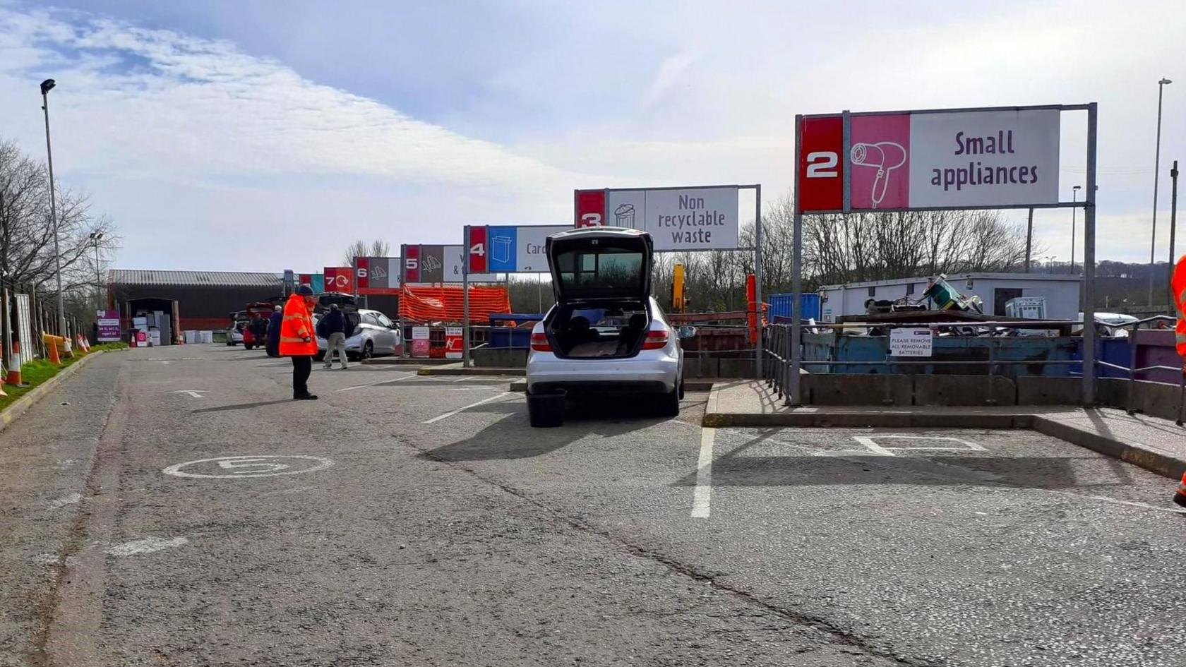 Daventry recycling centre, showing signs such as "small appliances" and "non-recyclable waste" alongside numbers for each skip. One person in orange hi-vis is watching people unload their cars while standing on a roadway. A white car near the camera has its boot open.