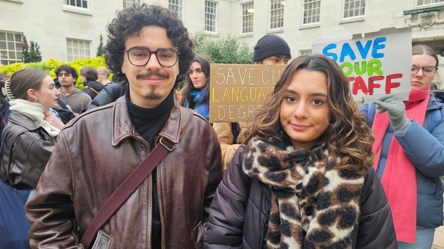 Markos Assiouras (right) and Anoushka Pathak (left). Markos has curly dark hair, glasses and is wearing maroon leather jacket. Anoushka has long dark hair and is wearing a black coat with a leopard-like print scarf. They are both stood in front of a crowd of people carrying protest signs. 