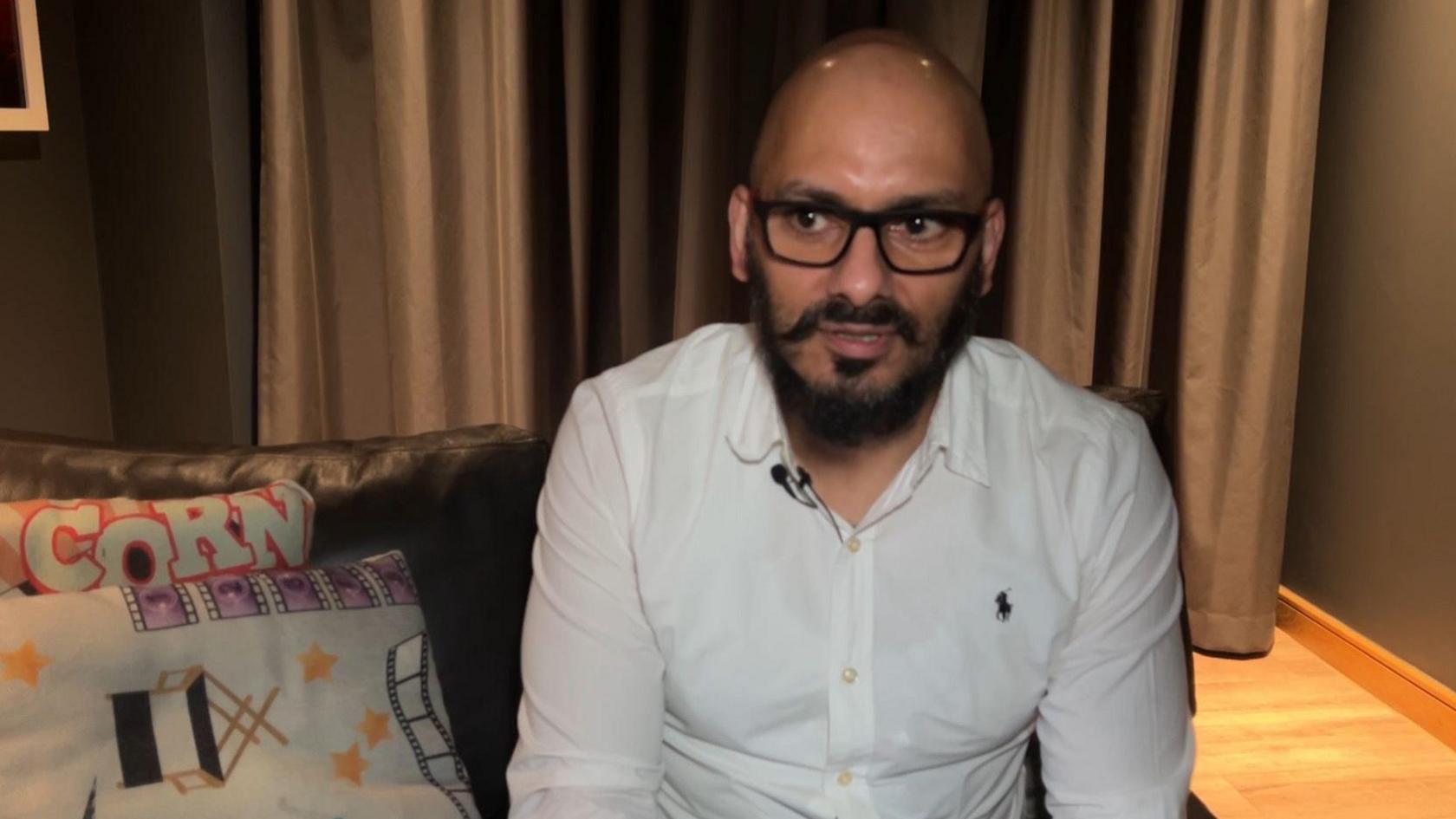 Vishal Duggal has a bald head, a black beard and moustache. He is wearing a white shirt and black glasses. He is sat on a leather sofa, and cushions behind him have a film theme, one with a caption saying 'Popcorn'. Behind him is a grey curtain. 