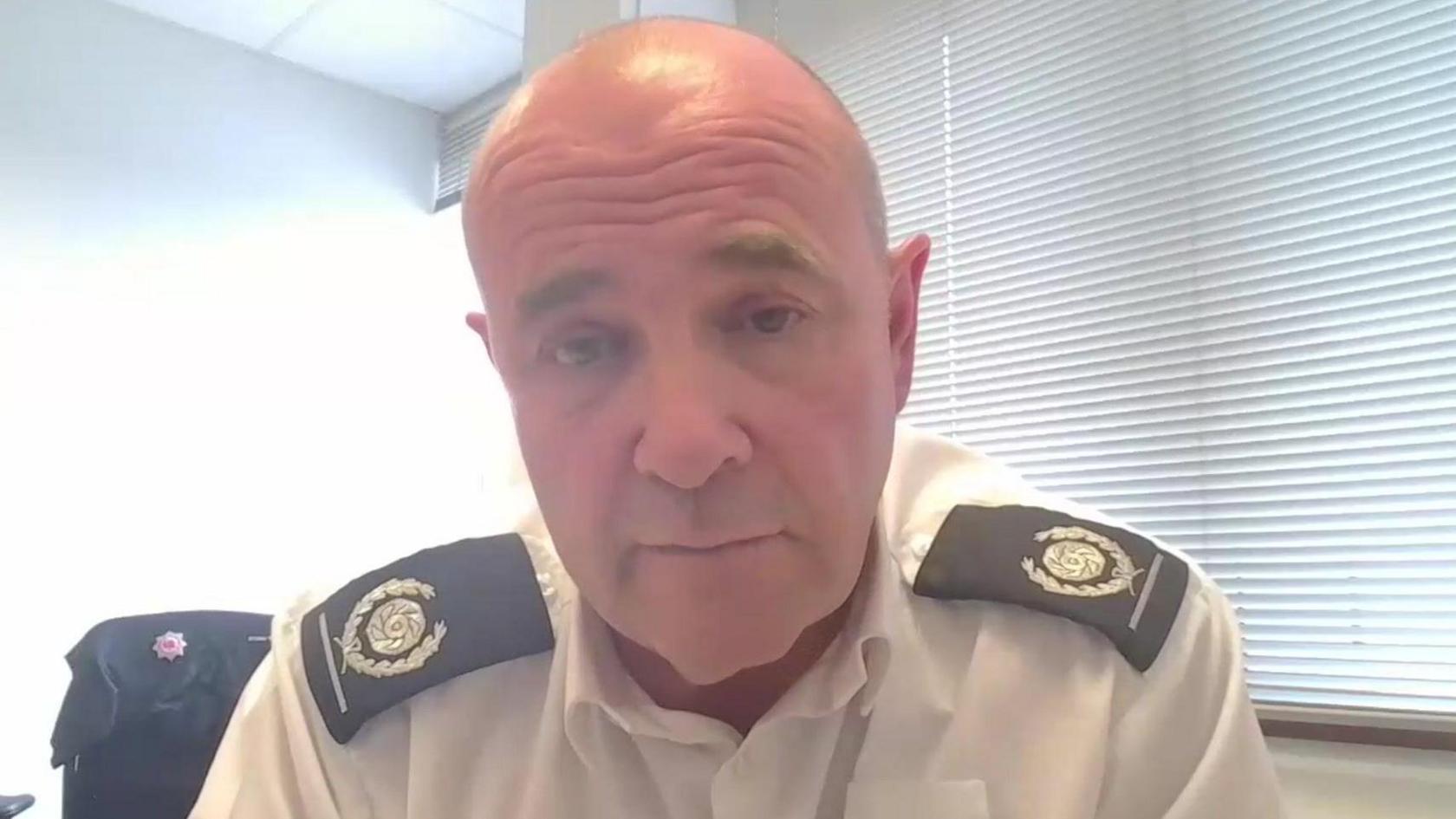 A close up picture of a man looking directly at the camera. He is wearing a white shirt with black fire service branding on his shoulders. There is a white blind in the background as well as a black jumper on a chair.
