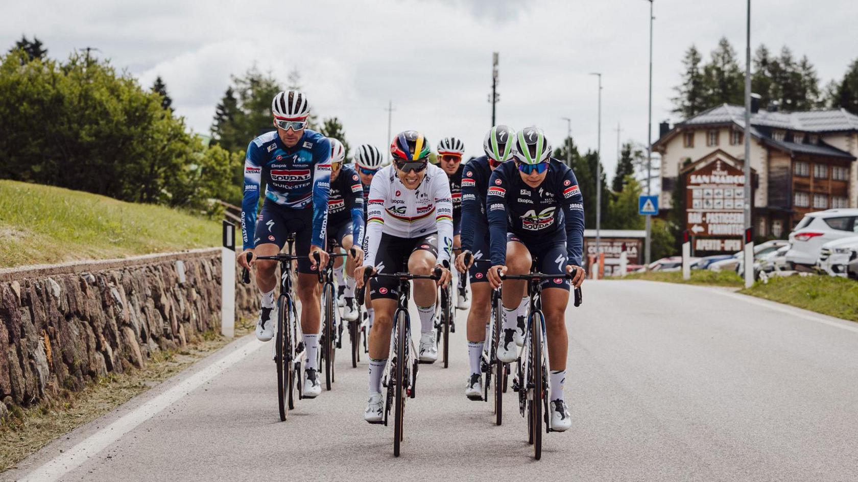 Seven cyclists, six wearing blue and one in the middle in white, are seen on a road with a chalet hotel in the background