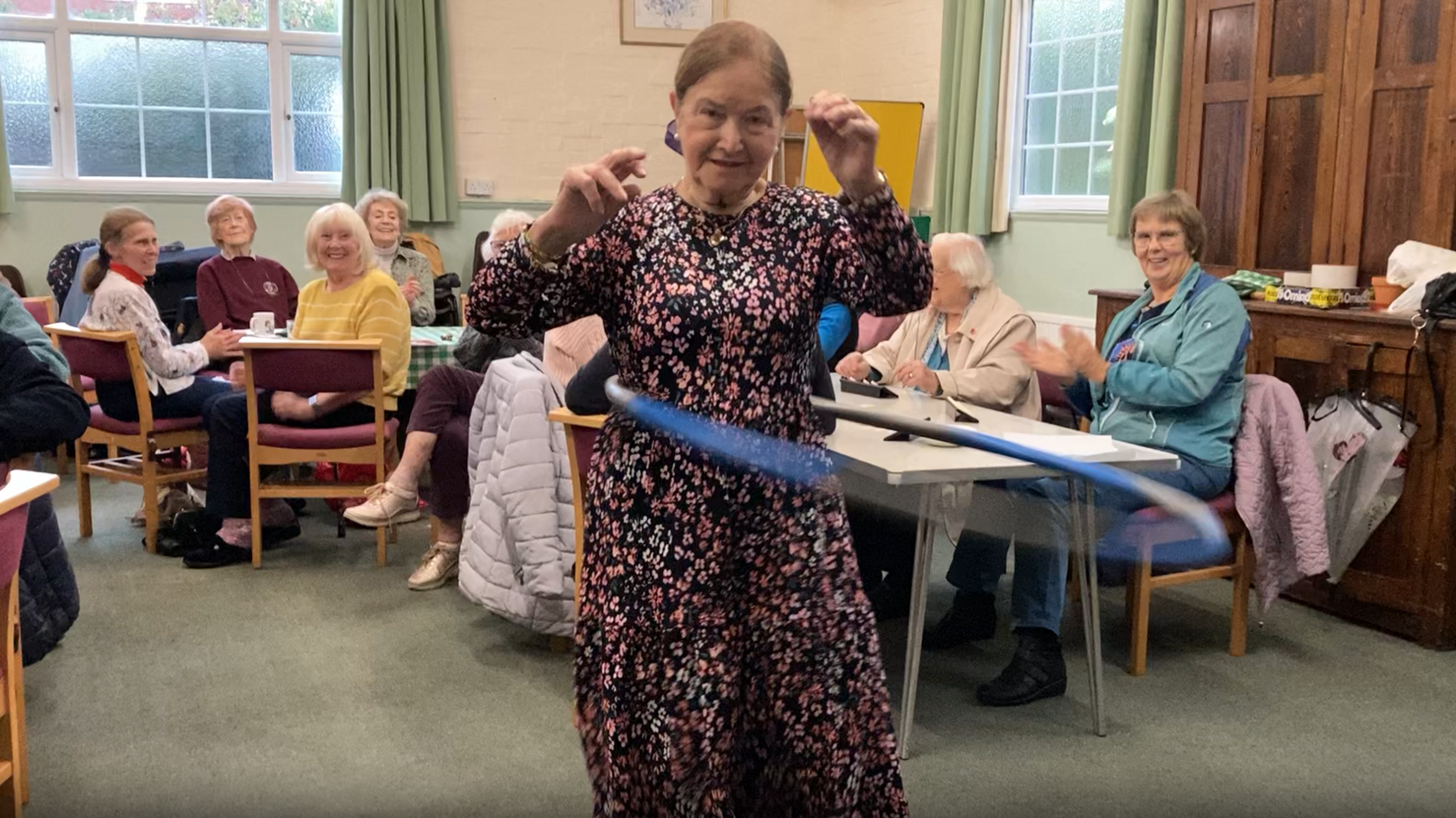 Nova Strange twirling a hula hoop around her while people watch seated in the background. She has tied-back brown hair and is wearing a long floral dress.
