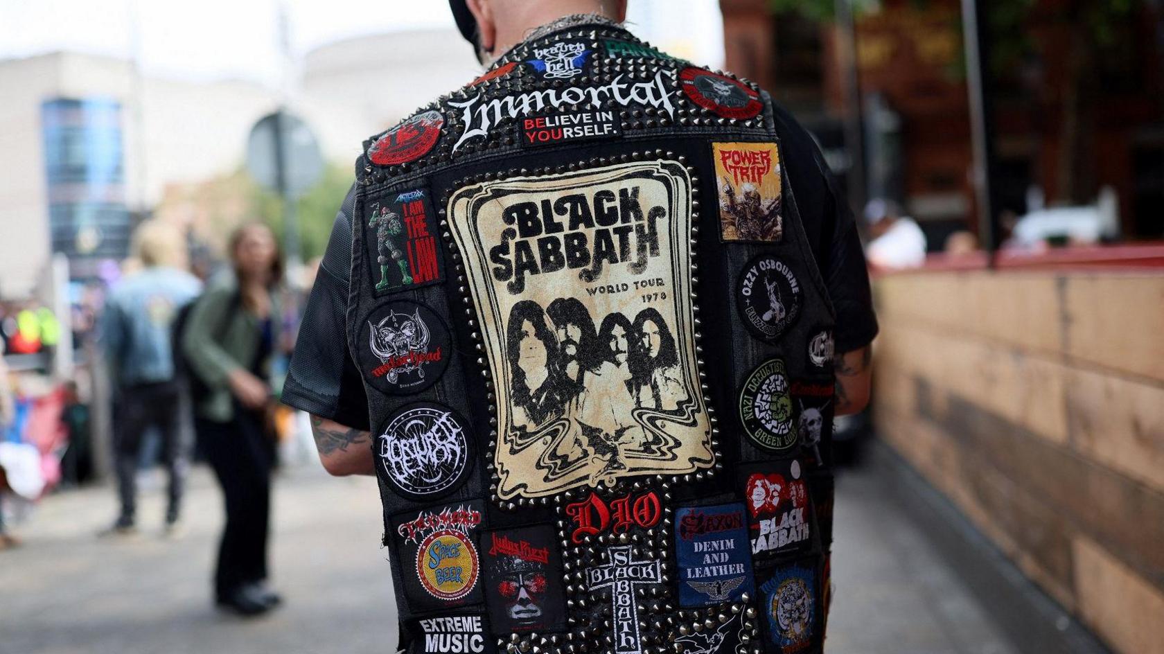 A man wearing a black denim jacket adorned with dozens of colourful patches depicting metal bands including Motorhead. There are metals studs near the collar. In the centre is a beige and black picture of the members of Black Sabbath