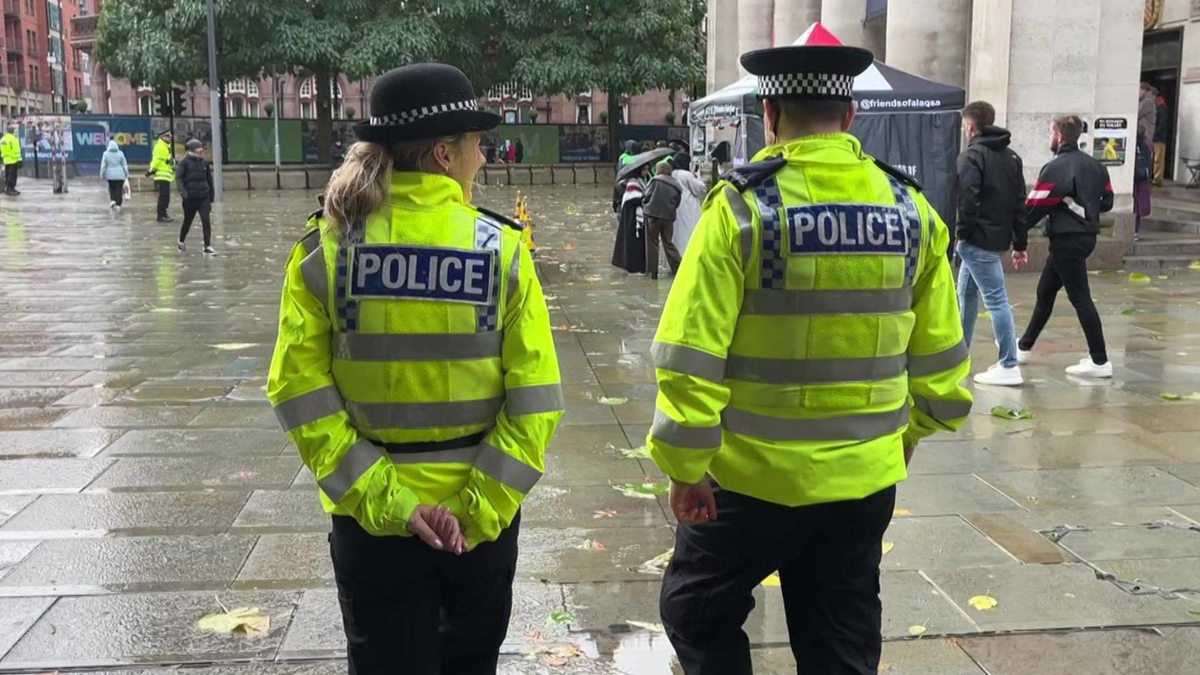 Two officers in police uniform including yellow reflective jackets, a woman with blonde hair and a man with short dark hair, walk across a paved square. 