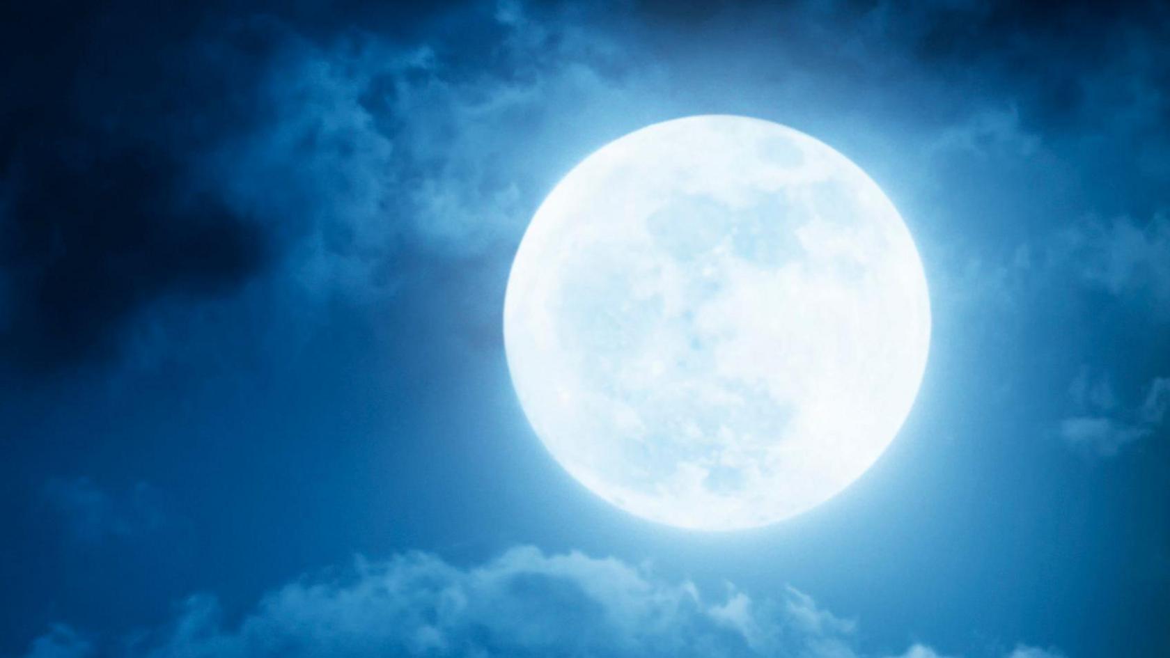 Large bright image of the Moon in the night sky with clouds around it.