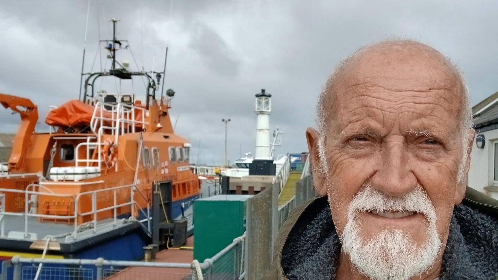 Derek Adams with short white hair and beard standing next to an orange lifeboat moored close to a white lighthouse on a black base.