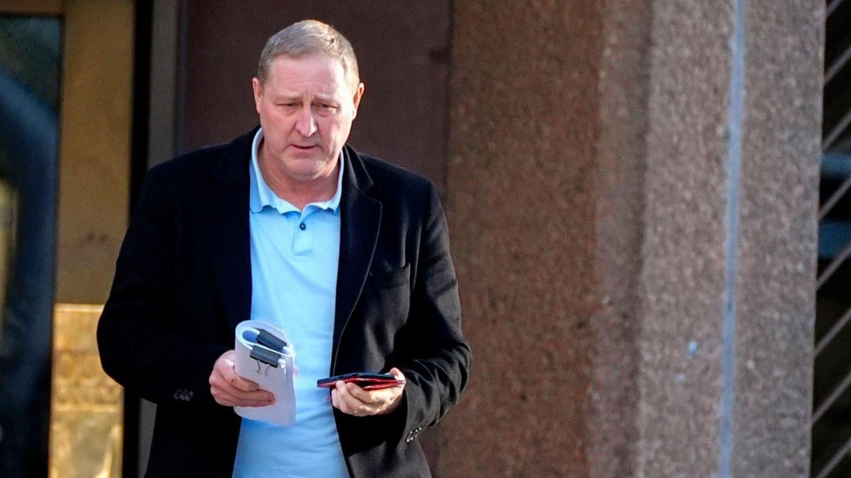 Paul Owen, with a black blazer over a blue shirt, walks away from Liverpool Crown Court holding a phone and a bundle of papers