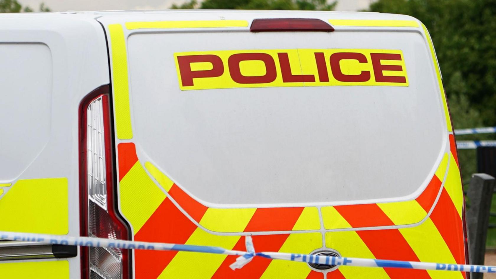 The back of a van with orange and yellow high vis stripes on it and police written in red across the rear door. Blue and white police cordon tape is stretched in front of the van and behind it.