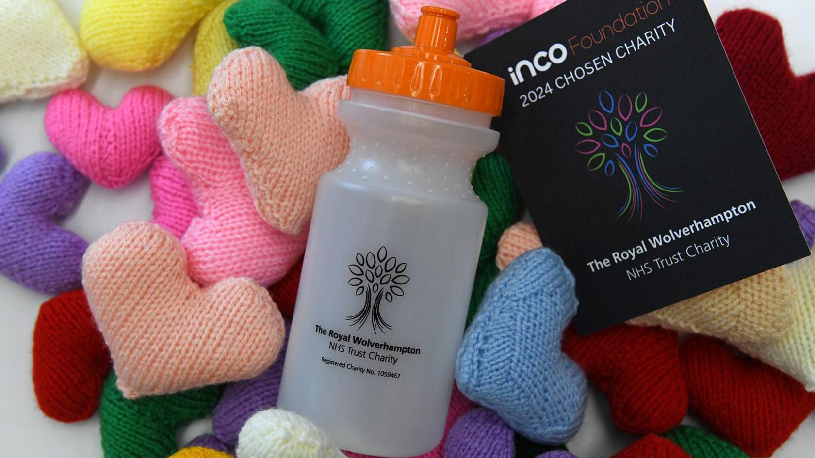 A white plastic bottle with an orange lid is on top of a pile of multi-coloured knitted hearts. It has a black logo on it that depicts a tree, and says "The Royal Wolverhampton NHS Trust Charity". Next to the bottle is a black leaflet with the same logo on the front.