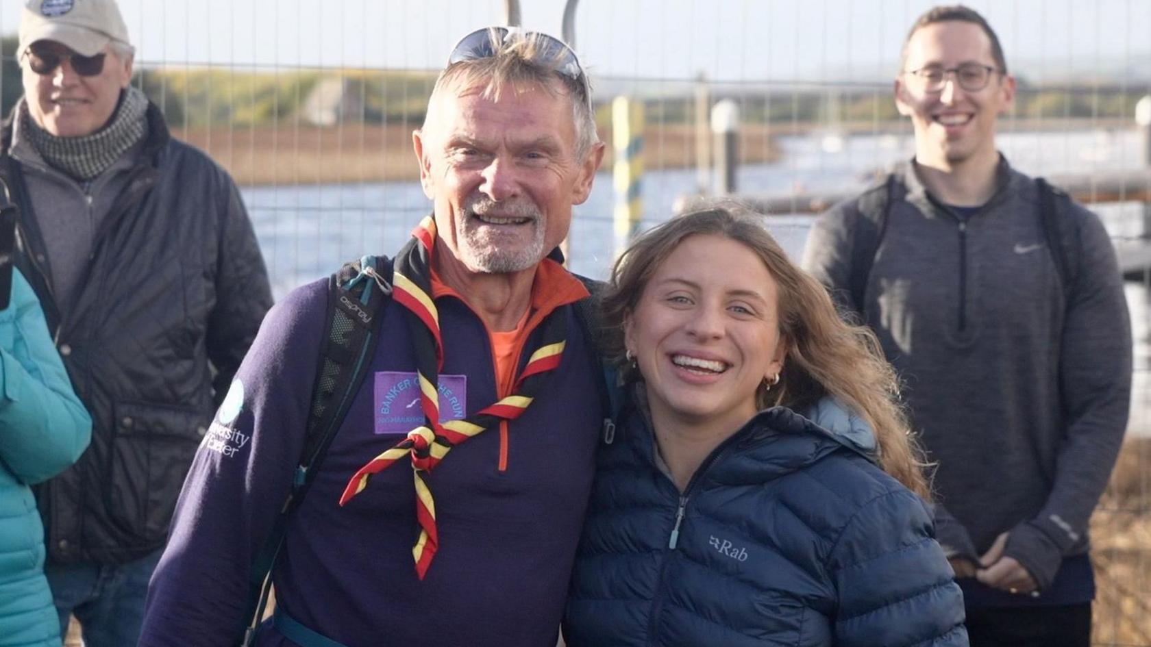 A man with embracing a woman from the side. They are shoulder-to-shoulder and looking towards the camera. The man has a goatee and has a neckerchief around his neck, and is wearing white-framed sunglasses on his head.