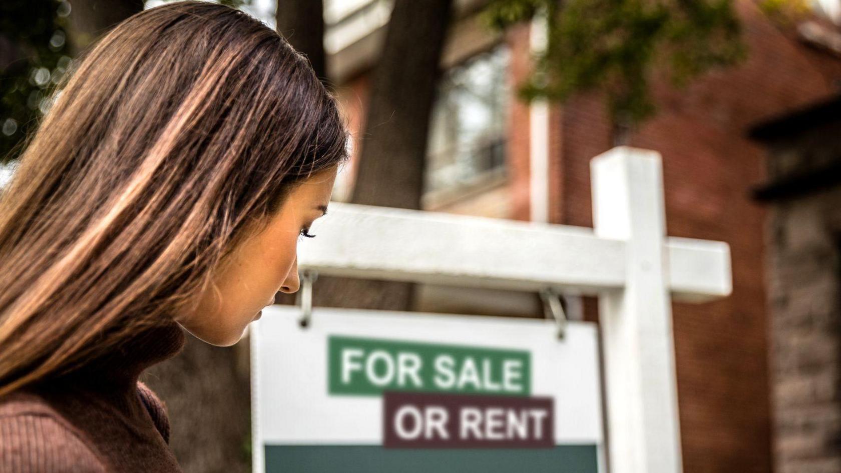 A woman with long dark hair and a brown top looking towards a house with a white sign in front of it that says "for sale or rent".