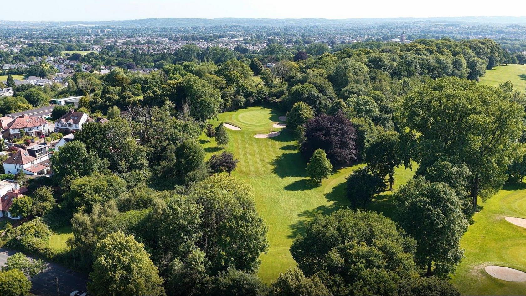 An aerial photo shows the seventh hole of the golf course, surrounded by trees. Several houses can be seen to the left and many properties can be seen in the distance.