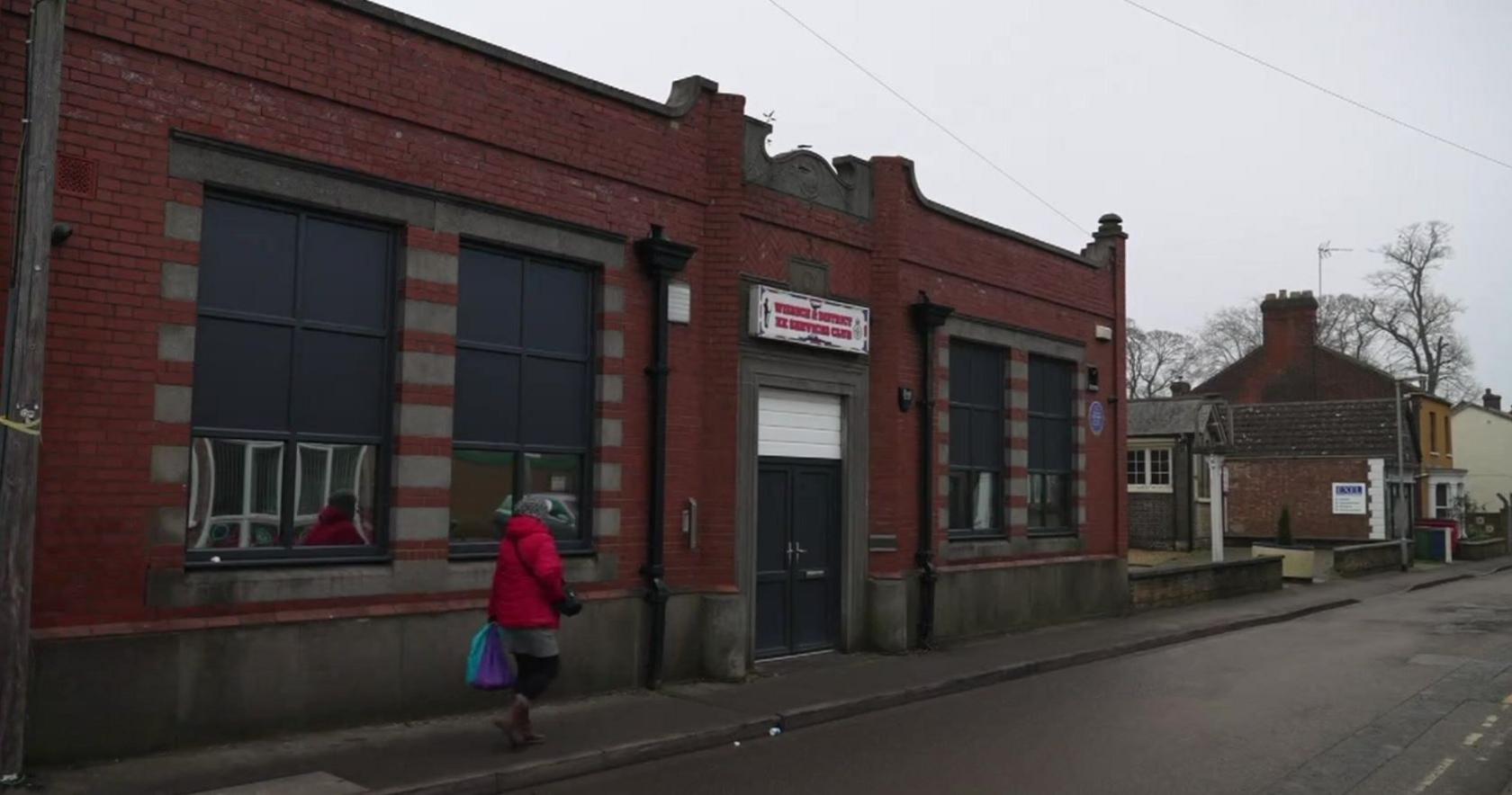 Wisbech former services club, a single-storey redbrick building with two large windows either side of a dark door which has a white sign above it with red writing displaying the building's name