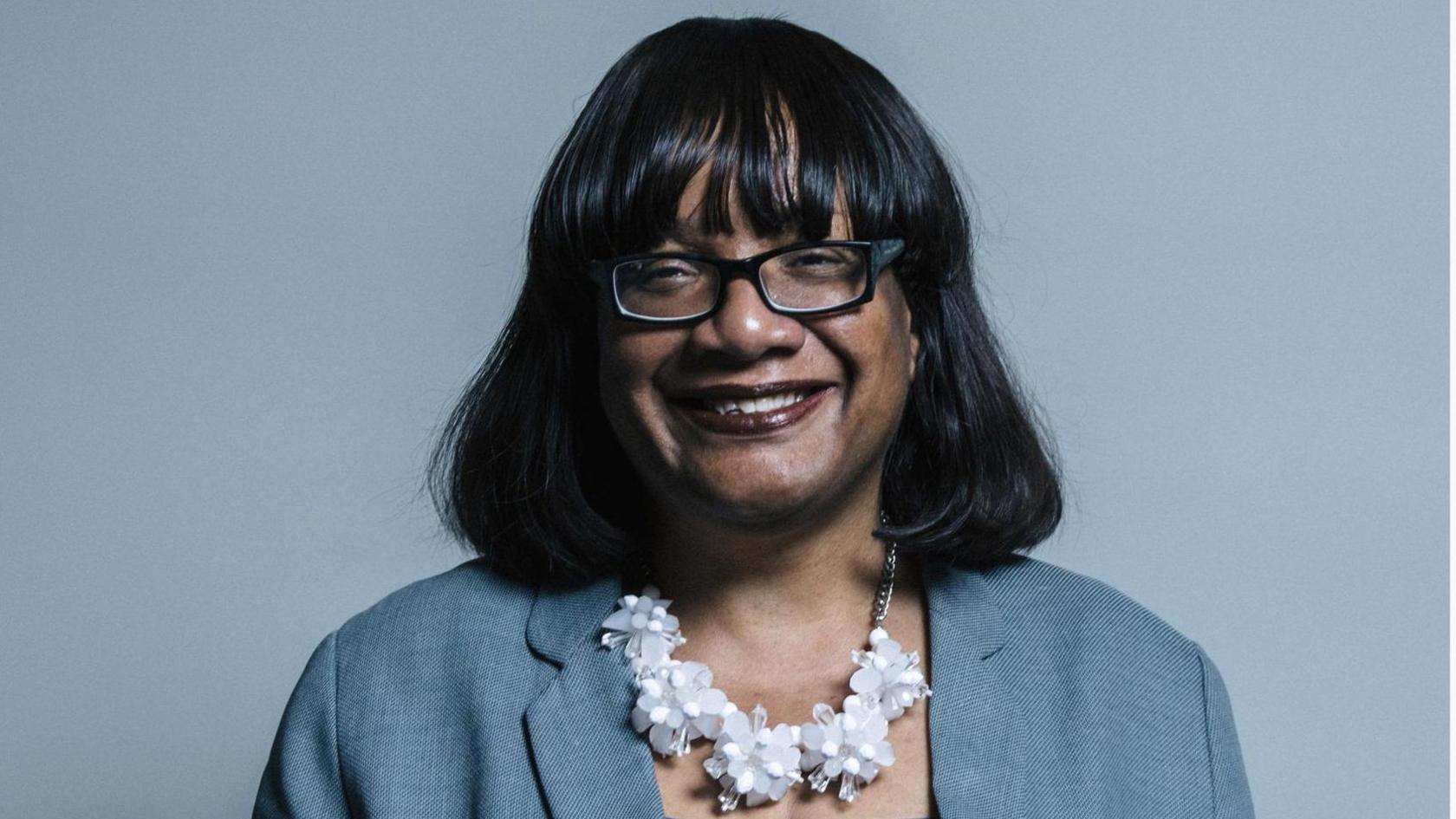 Diane Abbott smiling, is wearing a blue jacket and white flowery necklace and standing in front of a blue background