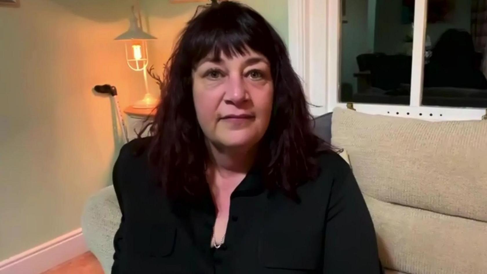 Hilary Bratton looking to camera during a BBC interview.  She has shoulder-length dark hair and is wearing a black blouse.
