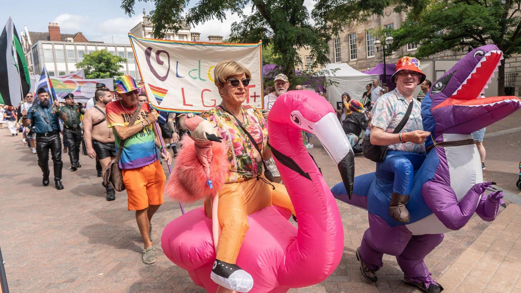 A person in an inflatable flamingo costume and another in an inflatable purple dinosaur walk together in a Pride Parade. The flamingo is bright pink, with a fake orange jockey legs and its rider is wearing a blonde wig and orange tropical print shirt and also holding a soft pink flamingo. The dinosaur jockey has fake blue legs and is wearing a blue pink and white patterned shirt and colourful bucket hat. Behind them others marching in a parade can be seen.