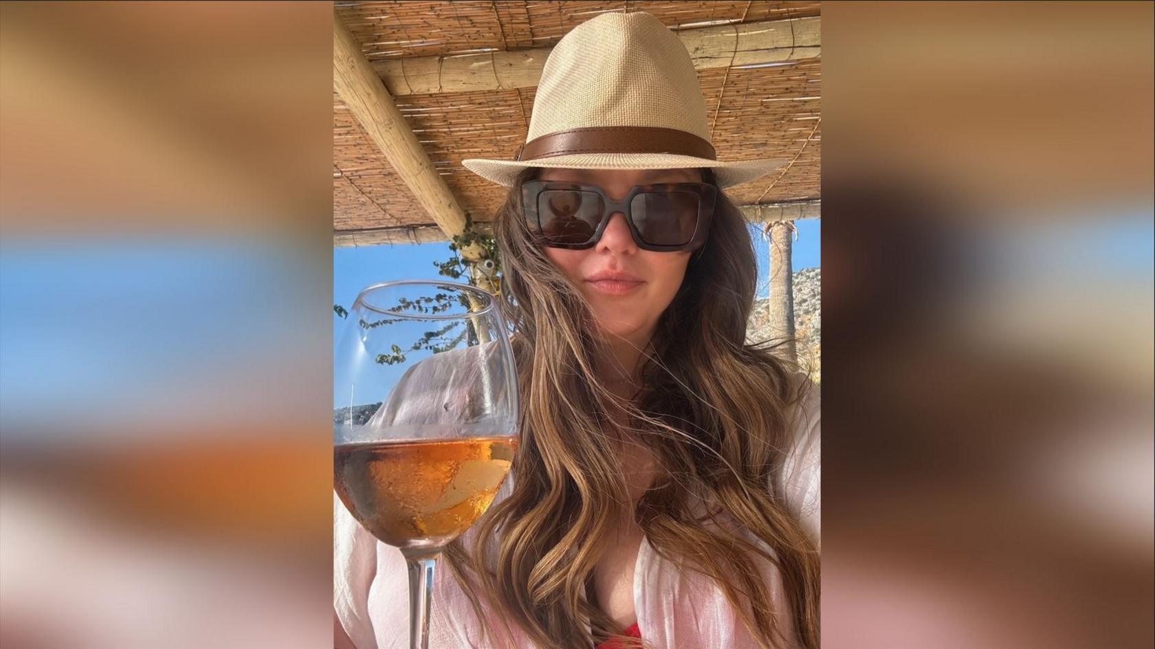 Melissa Clayton wearing large brown sunglasses, a cream hat, and white shirt. She has an orange drink in a wine glass in front of her. Picture taken on holiday.