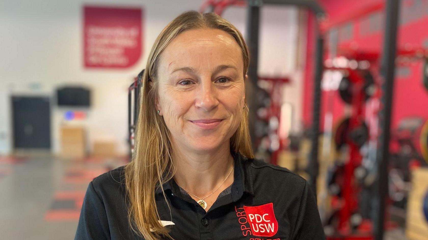 Dr Kate Williams has long brown hair. She is smiling at the camera, wearing a black polo shirt. There is red logo of University of South Wales staff on the right shoulder of her top. It is a head and shoulders shot of her.