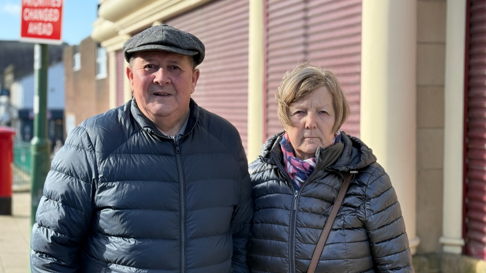 William Ringer, wearing a cap and a blue puffer jacket, stands next to Janice Ringer who has long brunette hair and is wearing a floral patterned scarf and a shiny black puffer jacket.