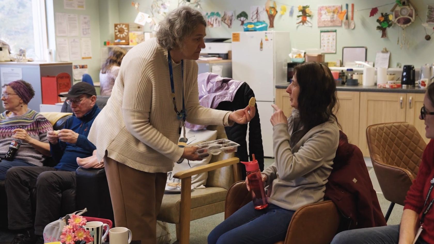 A shot from the film, showing older people sat around a room listening to music, whilst eating biscuits.