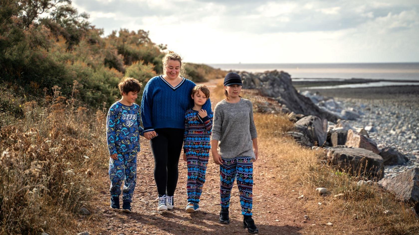 Sammiie wearing blue Converse shoes, black skinny jeans and a blue knitted jumper. She is walking alongside her three children on a coastal path next to the beach, which is full of rocks and shingles. It is a bright but slightly overcast day. All three of the boys are wearing blue patterned pyjamas. 