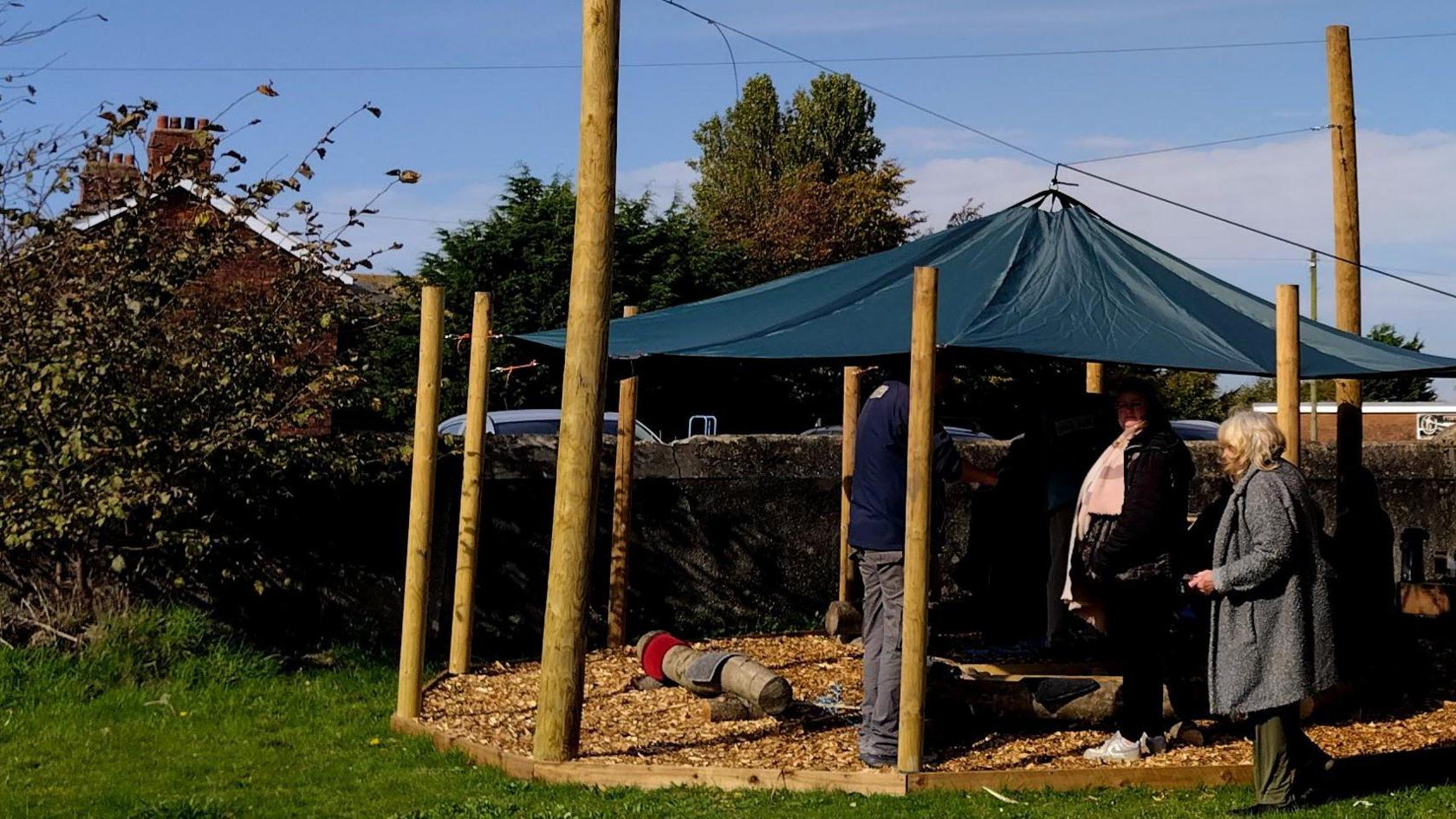 The new wildlife shelter in Preesall, Lancashire. Woodchip on the floor - a canopy in the centre of wooden posts - people gather around -  a house is in background