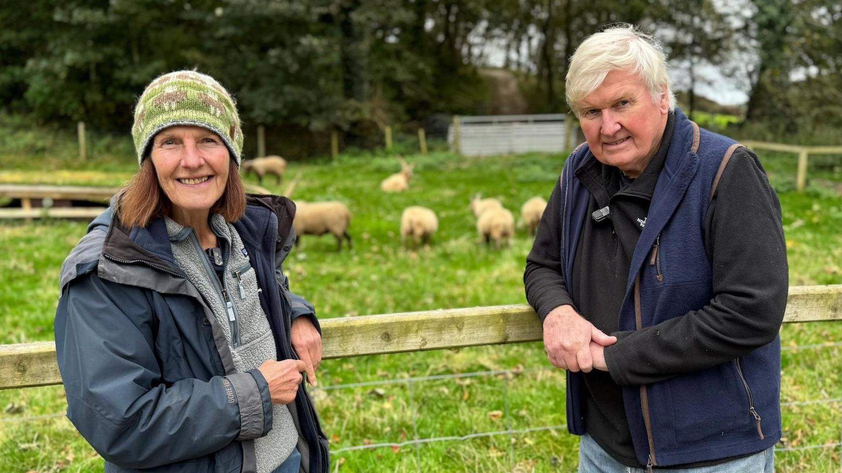 Jenny Shepherd is on the left leaning against a wooden fence, with her husband Rawdon standing to the right also leaning on the fence. They are wearing warm outdoor layers and Jenny is wearing a green woolly hat. In the field behind them can be seen a number of Loaghtan sheep grazing on the grass, with trees liing the back of the enclosure.