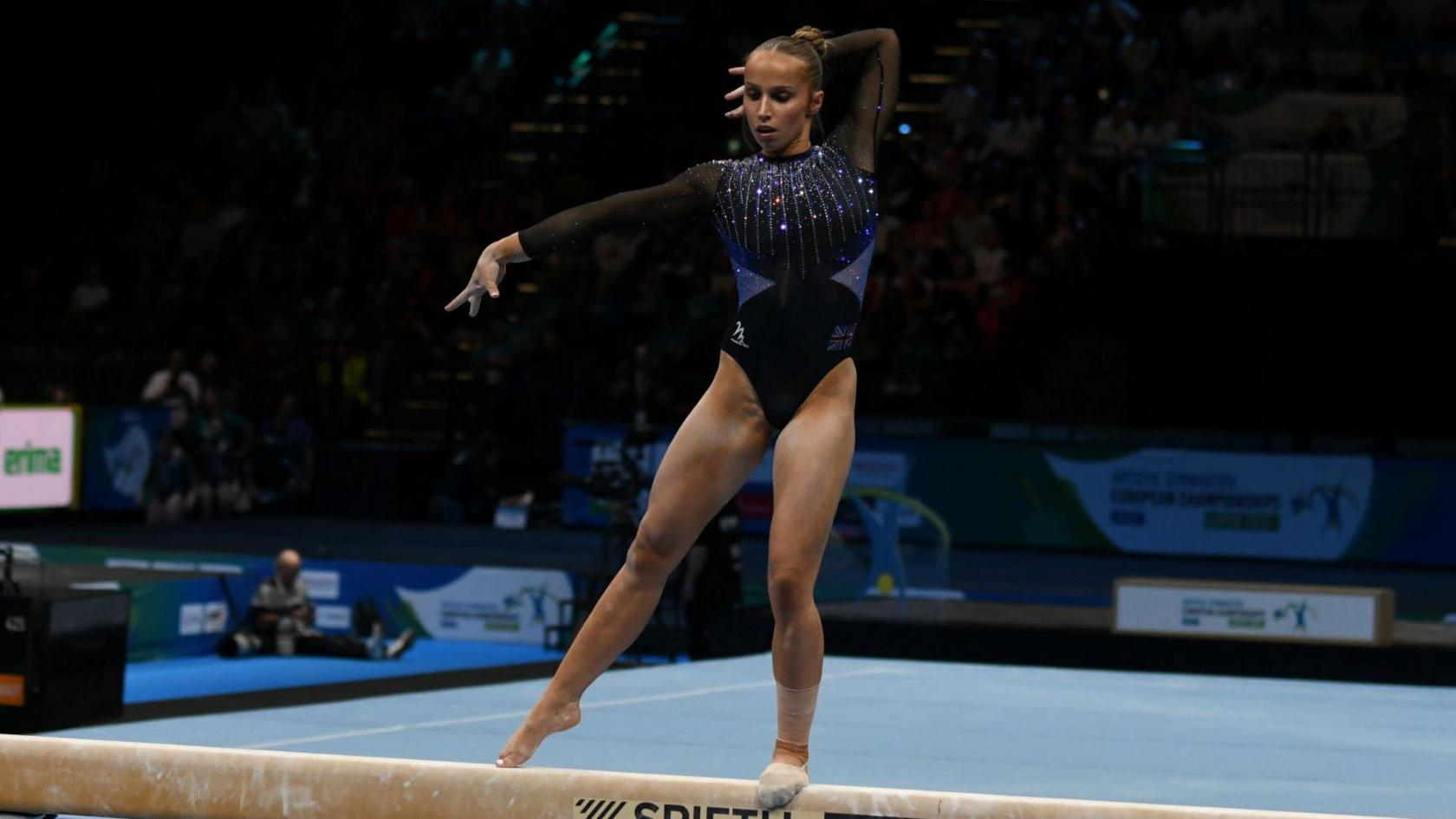 Emily Roper is wearing a sparkly blue leotard while standing on a bar during a competition. She has one leg slightly raised and her arms are pointed to her right.