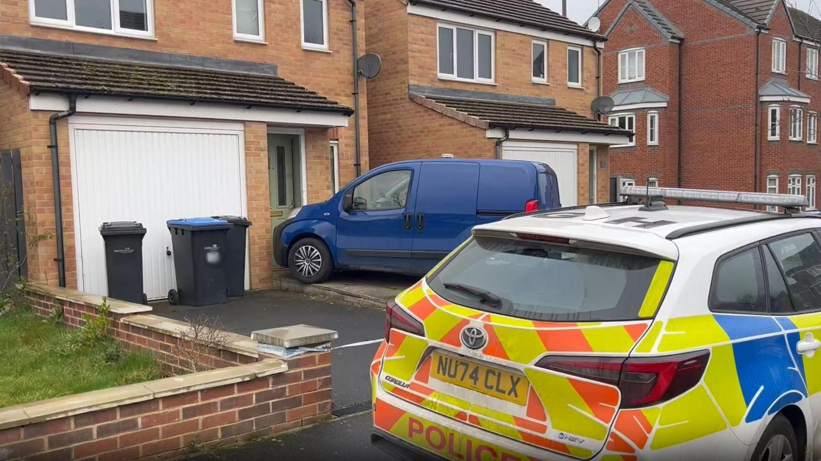 A police car parked outside a detached home. A small blue van is parked outside the property's front door, with its bumper almost touching the wall.