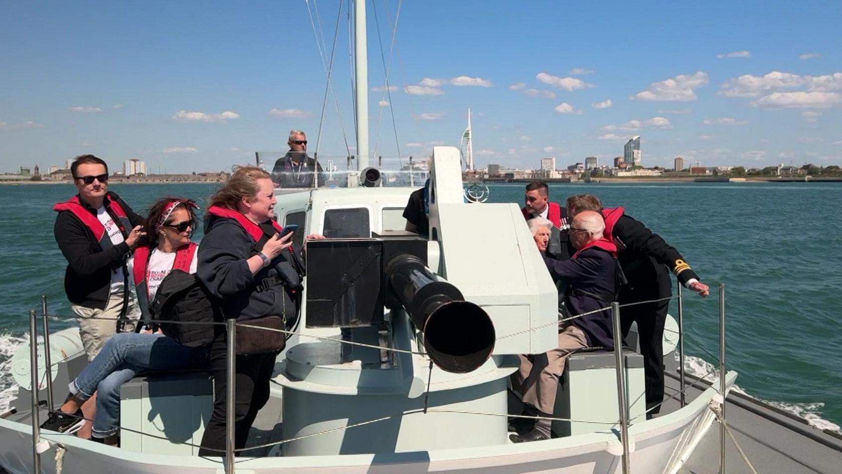 Looking at the boat from the bow, its large black gun barrel at the front. About eight people can be seen on the boat, including a couple of the veterans. Portsmouth harbour is in the distance.