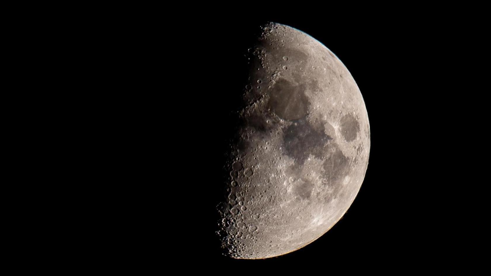 The moon fills this image against a black sky. Half of the satellite is in shadow, the other half is clearly visible with craters and dark spots