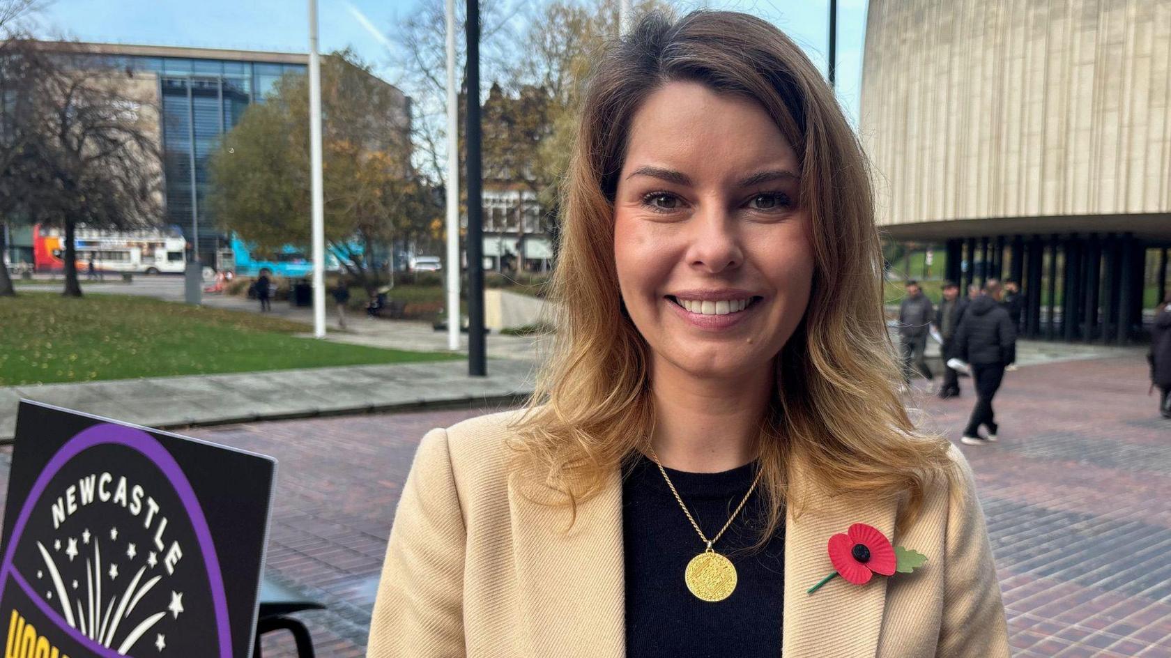 North East Mayor Kim McGuinness. She wears a cream coat with a poppy, and is standing in front of the Newcastle Civic Centre smiling at the camera. To her left the top of the sign for the HogmanHoway 5K is slightly visible.