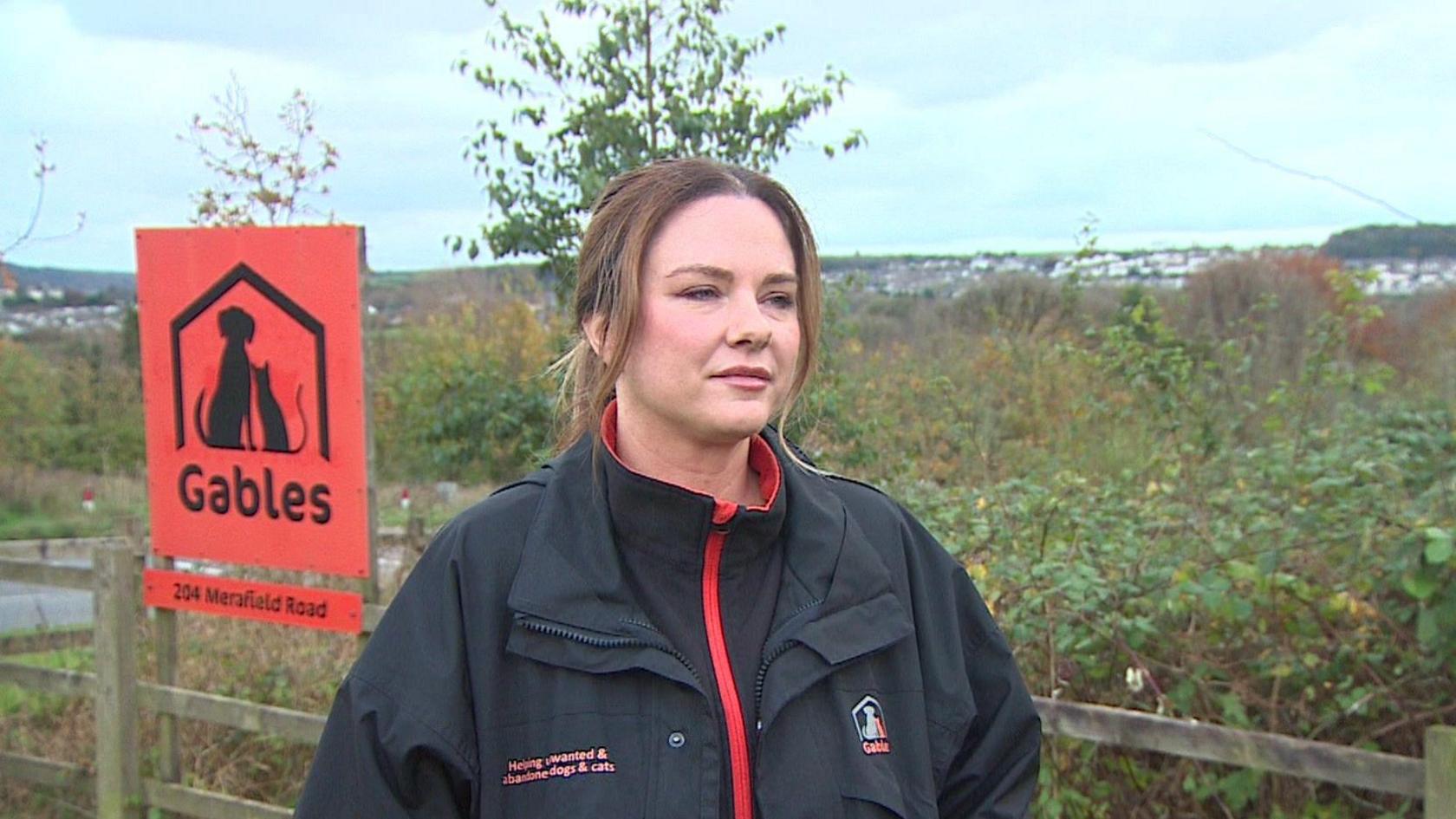 Claire Sparkes looking to right wearing a black coat with a red zip and red writing on the left. There is a logo on the right of the coat. There is an orange sign on the left behind her with a silhouette of a dog and cat under a roof. Under the logo is the words 'Gables' and '204 Merafield Road.' Behind the sign is the plot of land which has green bushes and a wooden fence around it 