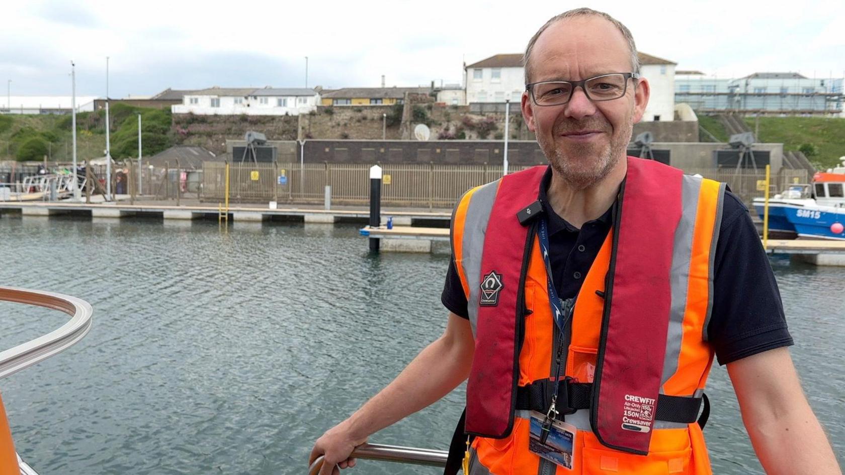 Tom Willis, a middle-aged man with glasses, stands on the deck of the boat smiling at the camera wearing a lifejacket and hi-vis.