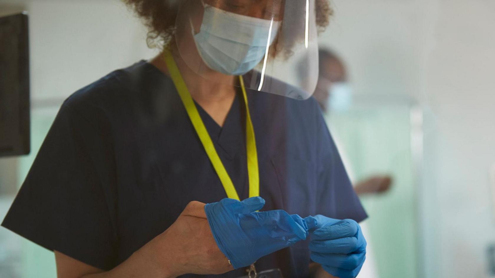 Medical professional wearing dark blue scrubs with a yellow lanyard. She is wearing a face covering including visor and in the process of putting on protective gloves