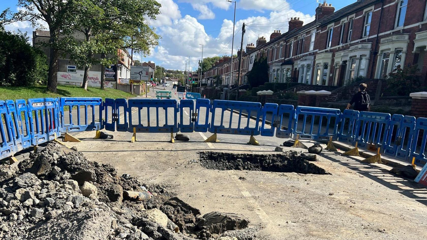 A view of the hole, dug out as a square in the tarmac. A large pile of debris from it to the left. Blue barriers all around the hole in the middle of the road. Red brick terraced houses line the right of the road, trees to the left. A sunny day.