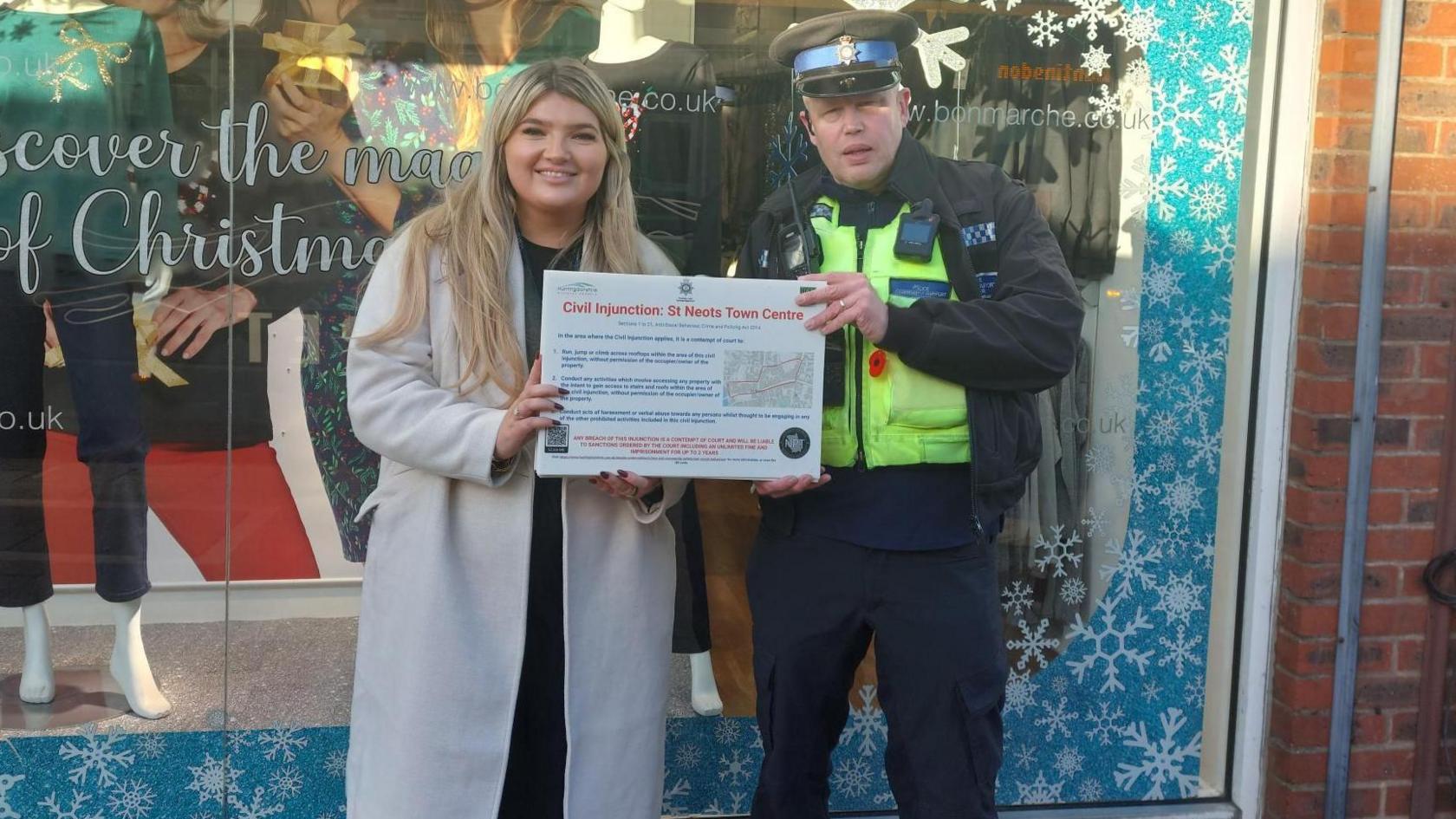 Georgia Patterson, community safety partnership delivery officer and PCSO Daniel Grant, standing outside a shop front holding a sign. Georgia is to the left and wearing a light coat, and has long fair hair, PCSO Daniel Grant, is wearing full police uniform, with a high viz jacket. 