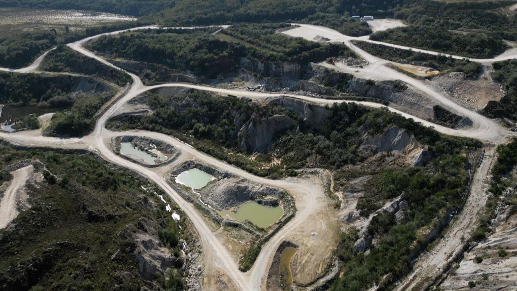 Ariel view of Trelavour pit. There are green areas amongst grey stone paths leading to three pits. The pit on the left has blue coloured water inside, the one in the middle has blue water and the one on the right has green water. They are surrounded by cliff faces covered with trees.