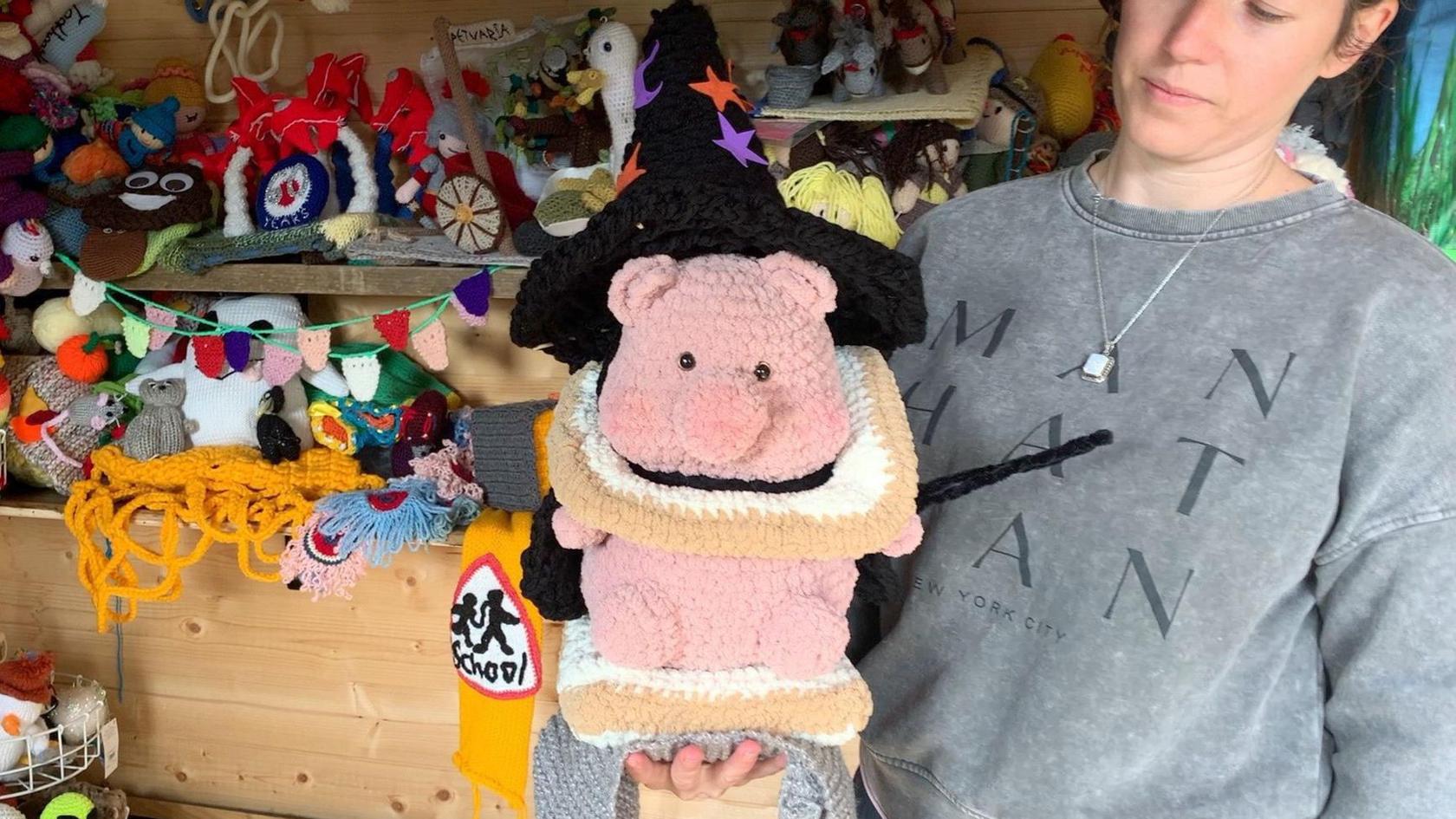 Alison Davis holds "Kevin Bacon", a knitted pink pig, which is encased in two knitted slices of bread and wearing a black witch's hat. In the background are shelves full of more knitted toppers.