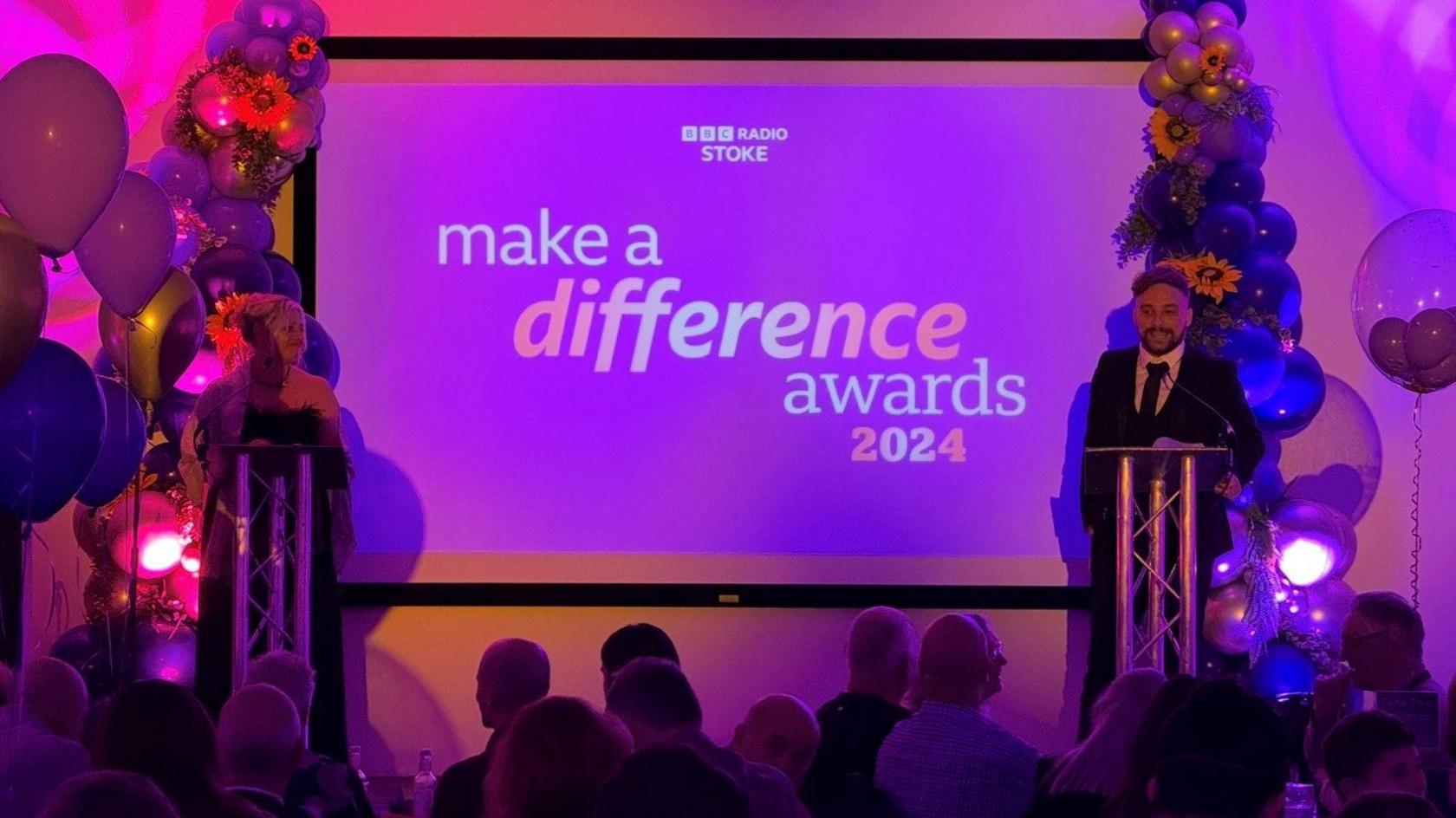 Jodie Looker and Lee Blakeman are stood at podiums either side of a stage which is illuminated with pink lighting, with the a purple backdrop which has writing on it which reads BBC Radio Stoke Make a Difference Awards 2024.