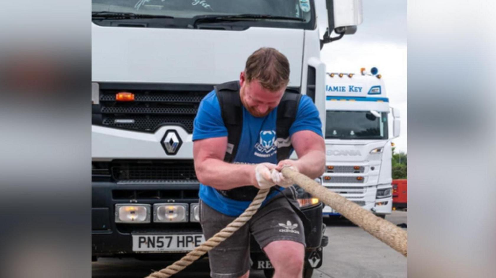 A man wearing shorts and a t-shirt pulls a lorry using a rope. He is visibly straining with the effort.