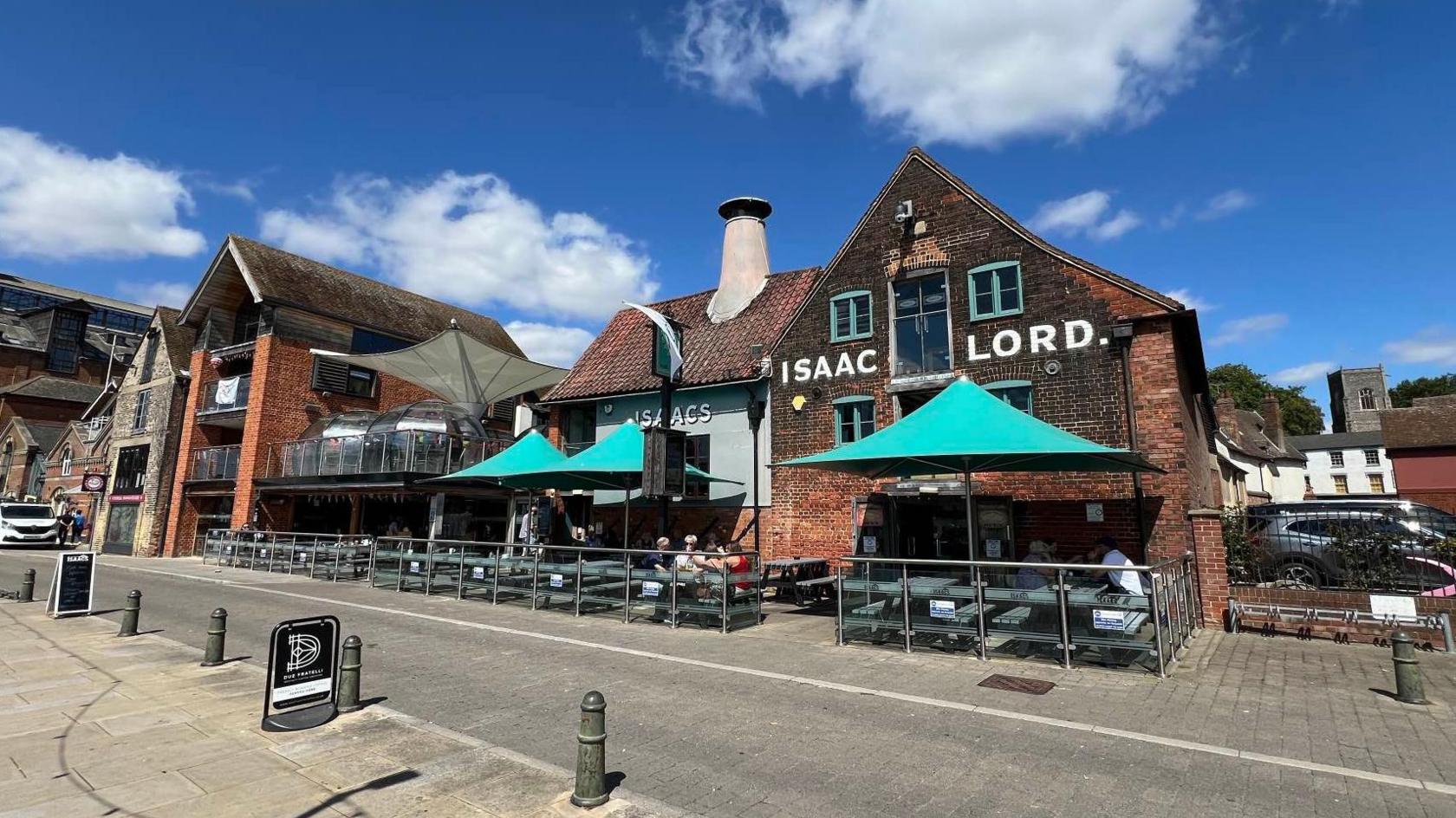 A pub with the sign the Isaac Lord painted onto its old red bricks, with large sea green parasols over benches and tables with people sitting at them