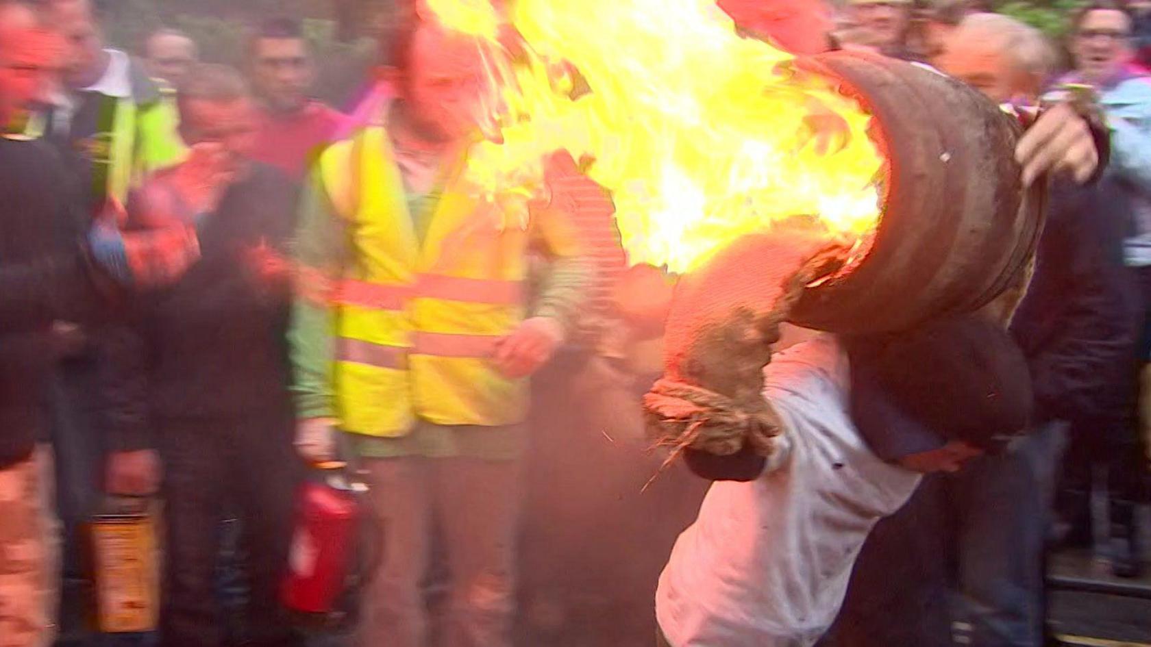 A young person carrying a flaming tar barrel on their back. There is a person stood behind him with a yellow hi-vis and a fire extinguisher in his left hand. There is a crowd of people looking at him.