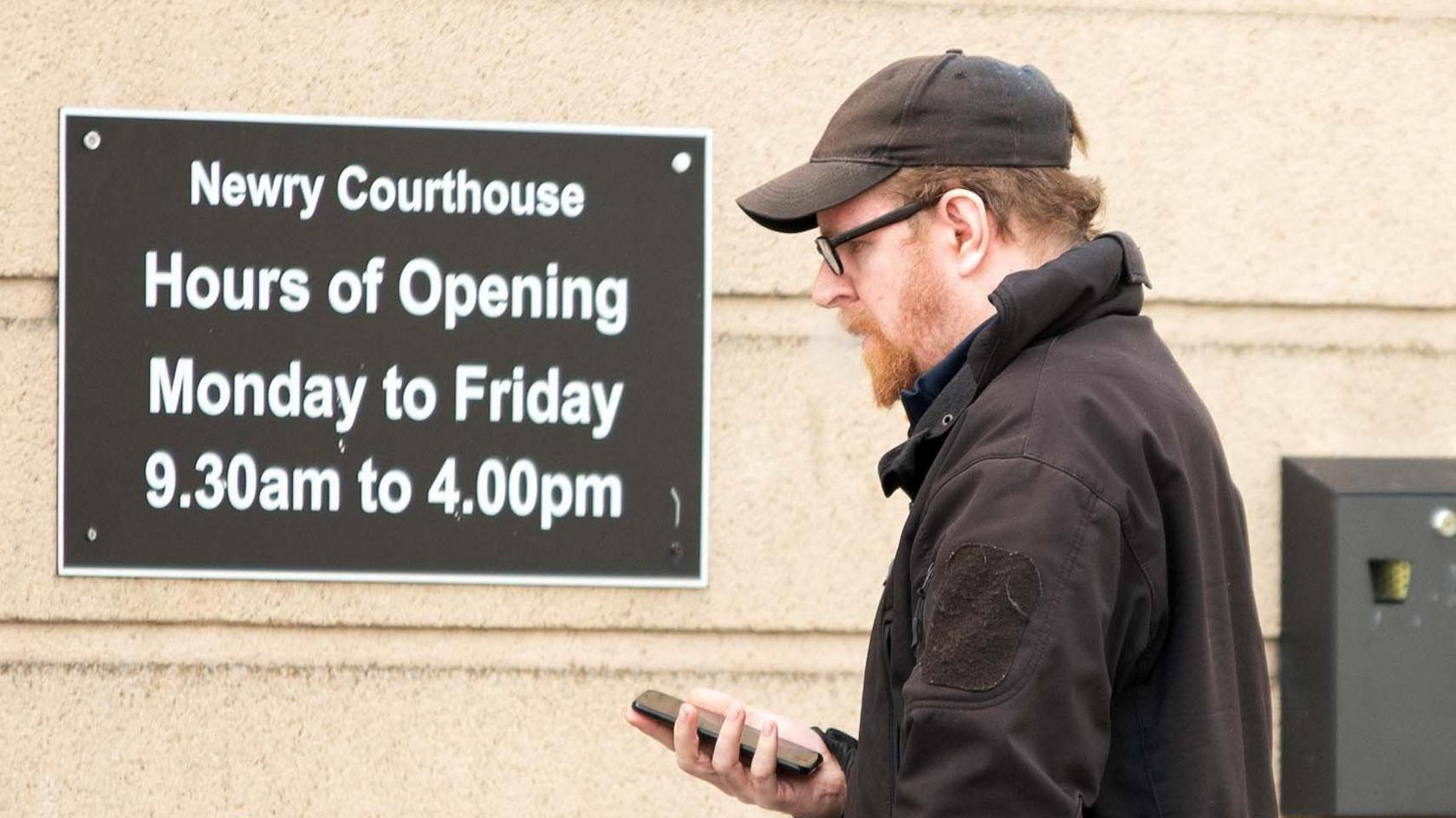 Chris Fulton is pictured holding his phone and wearing a black cap. He is wearing black framed glasses and a black coat. He is walking past a sign that says Newry Courthouse, Hours of opening, Monday to Friday, 9.30am to 4.00pm.