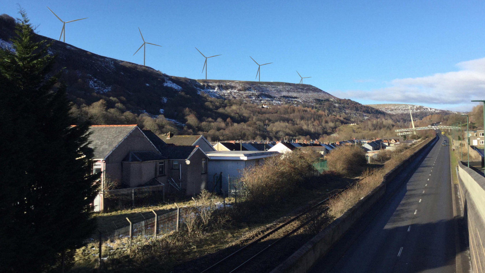 A mock up of a road and railway line next to a row of terraced houses in front of some steep hills in the background. On the stills there are five impressions of wind turbines 