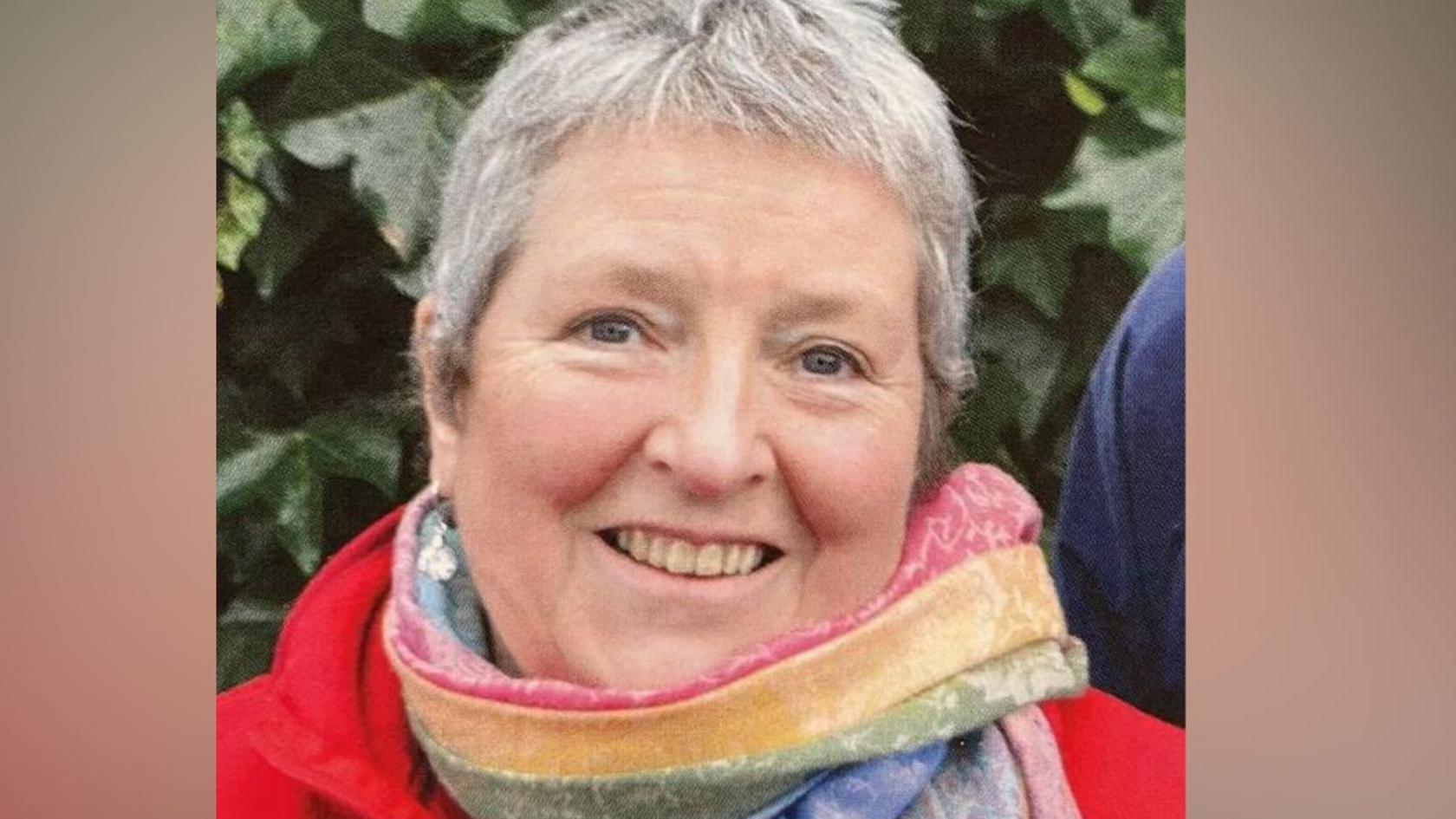 Councillor Judith Derbyshire. She has short grey hair and is wearing a red coat and colourful striped scarf. She is outdoors, stood in front of a leafy bush.