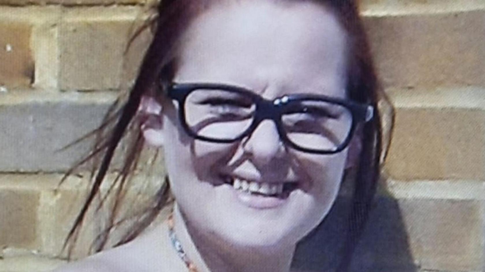 A woman in dark glasses with dyed auburn hair smiling. She is standing in front of a brick wall.